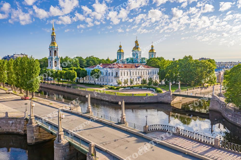 На фотографии изображен Николо-Богоявленский морской собор Петербурга с его золотыми куполами и высокой колокольней, окруженный зелеными насаждениями и каналами. Каналы, пересекающиеся с мостами, придают сцене очарование классического Петербурга. Яркий летний день подчеркивает гармонию архитектуры и природы.